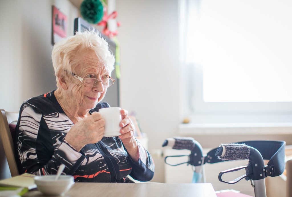 Seniorin Rosemarie John lächelt zufrieden mit Kaffee in der Hand am Tisch im Seniorenheim.