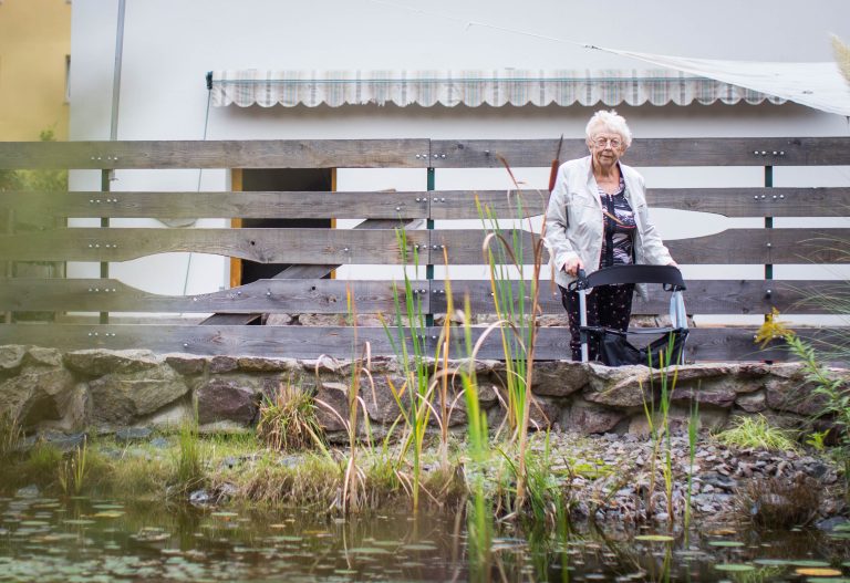 Seniorin Rosemarie John geht mit ihrem Rollator vor dem Seniorenheim spazieren im Park.