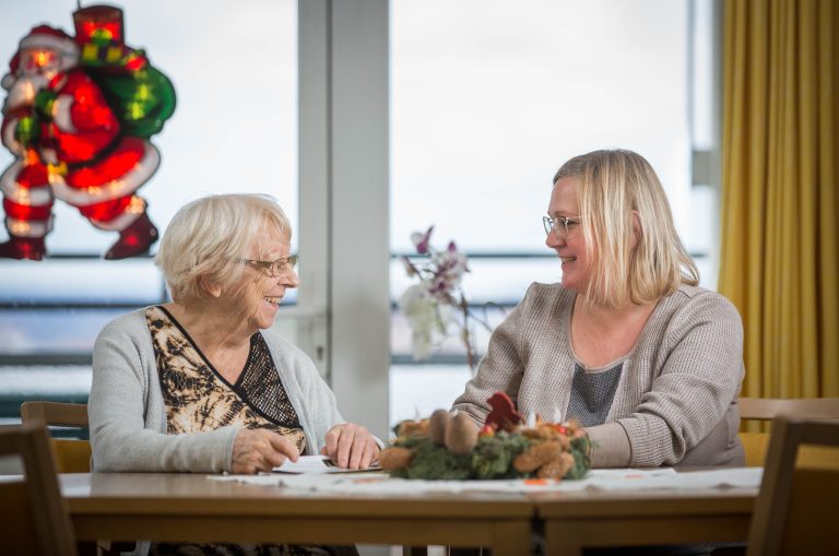 Ines Karsten sitzt lachend im Gespräch mit Rosemarie John im Pflegeheim am Tisch.