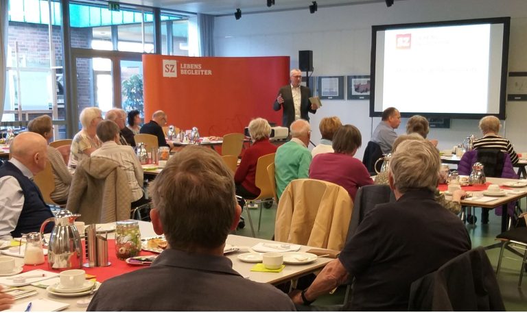 Im Veranstaltungssaal sitzen viele Gäste bei Kaffee und Kuchen und hören dem Moderator vom Gesundheitsvortrag zu.