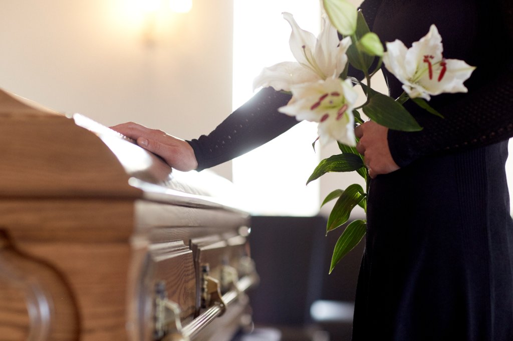 Frau in schwarz mit Lilien im Arm legt eine Hand auf den Holzsarg.