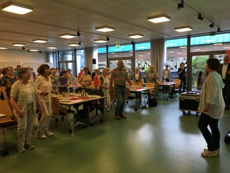Im komplett gefüllten Saal führt eine Therapeutin Übungen vor und das Publikum macht stehend mit.