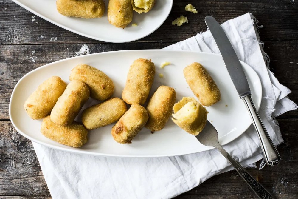 Knusprige Kartoffelkroketten liegen auf einem Servierteller und in einer steckt eine Gabe drin.