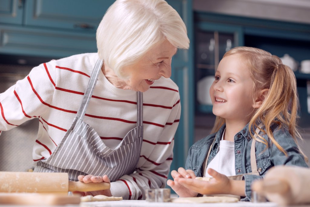 Eine weißhaarige Seniorin mit Schürze und ein lächelndes Mädchen mit Zöpfen rollen gemeinsam den Teig aus. Foto: AdobeStock/zinkevych