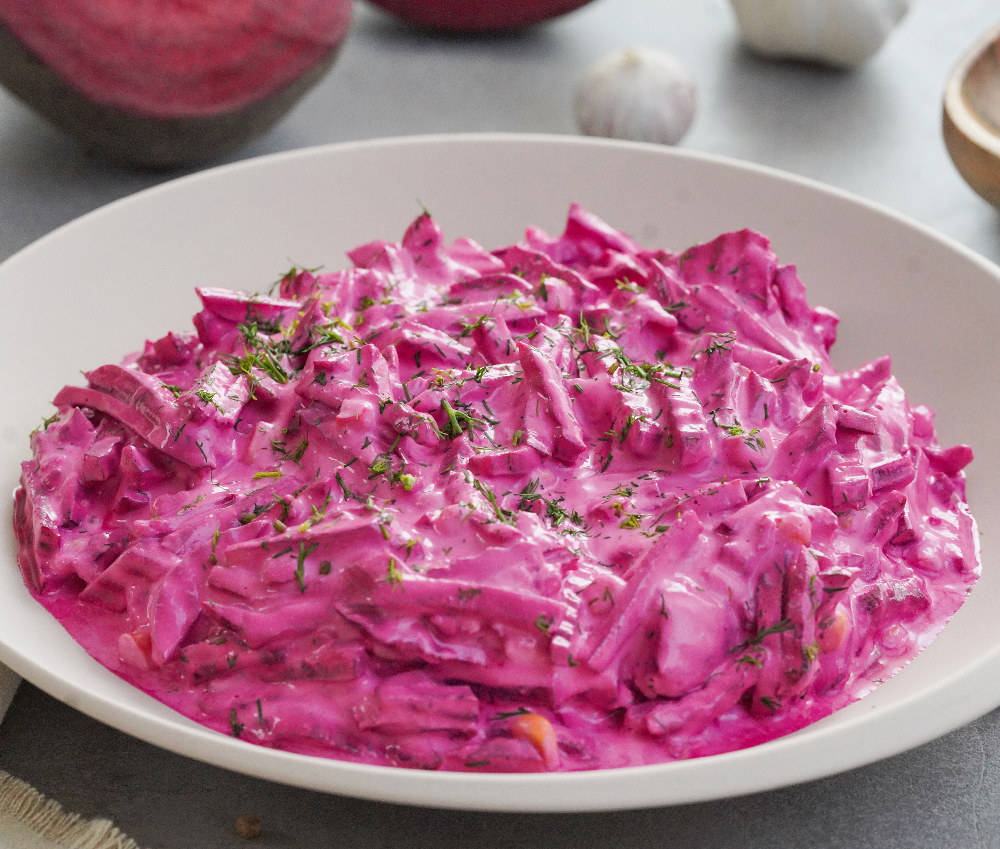 In einem weißen, tiefen Porzellanteller ist ein lila Rote-Beete-Salat angerichtet. Dahinter liegt Knoblauch und eine rote Zwiebel.